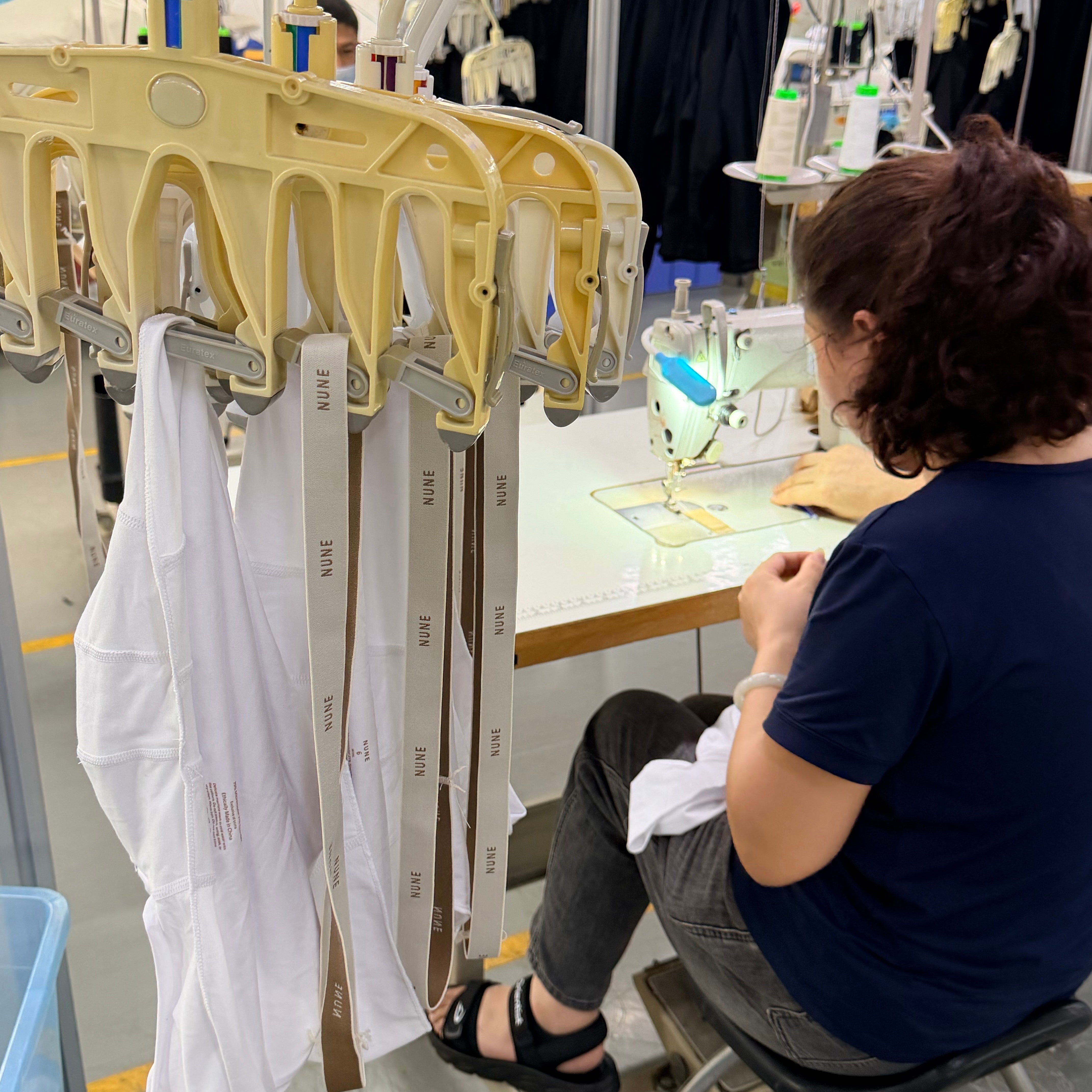 A person seated at a sewing machine in a garment workshop, working with white fabric pieces hanging on a yellow garment rack. Industrial sewing equipment and racks are visible in the background.