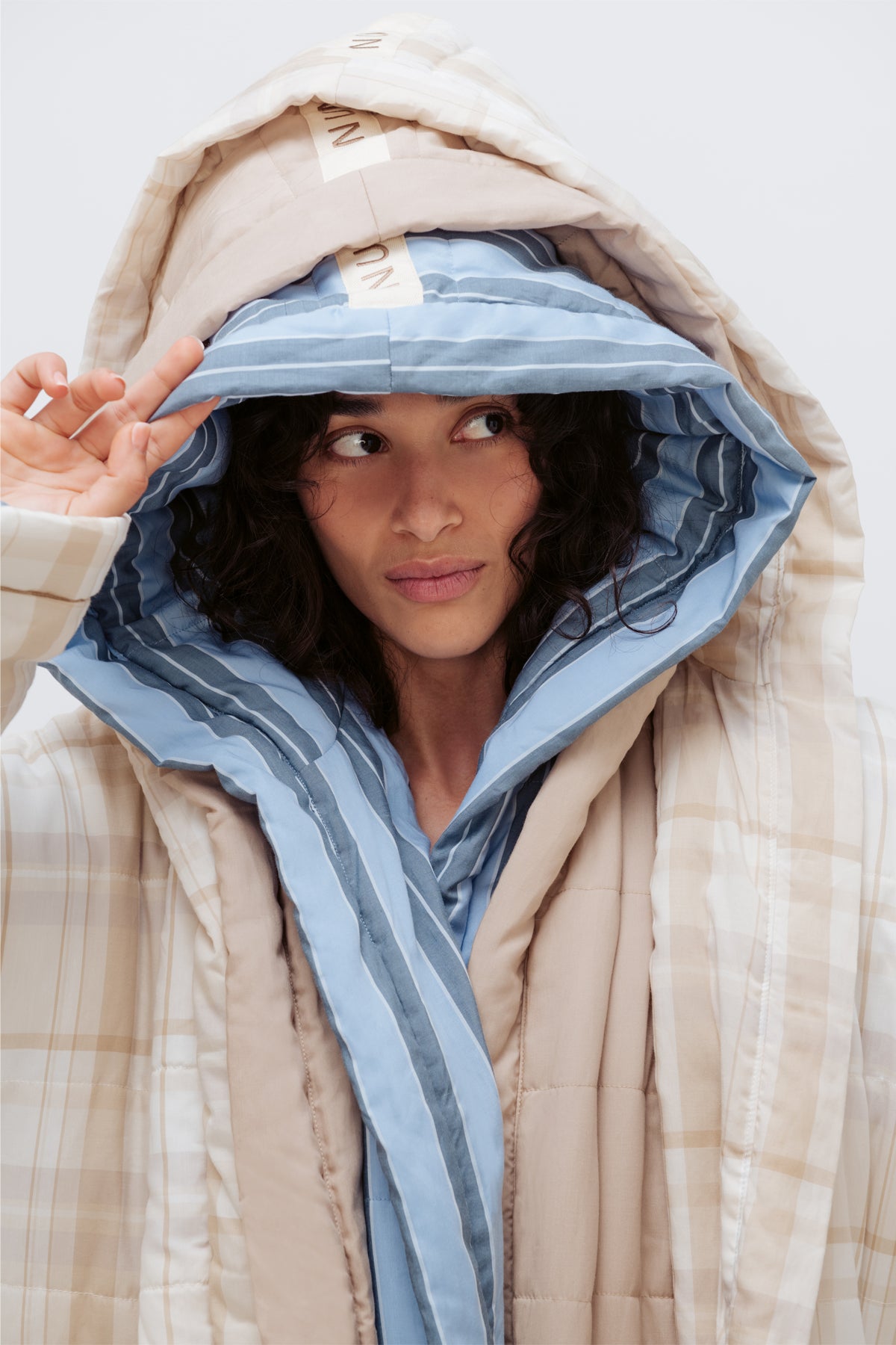 A close-up of a model wearing a beige quilted puffer jacket with a striped hood, layered over a light blue striped shirt. The model is adjusting the hood of the jacket against a plain light gray background.
