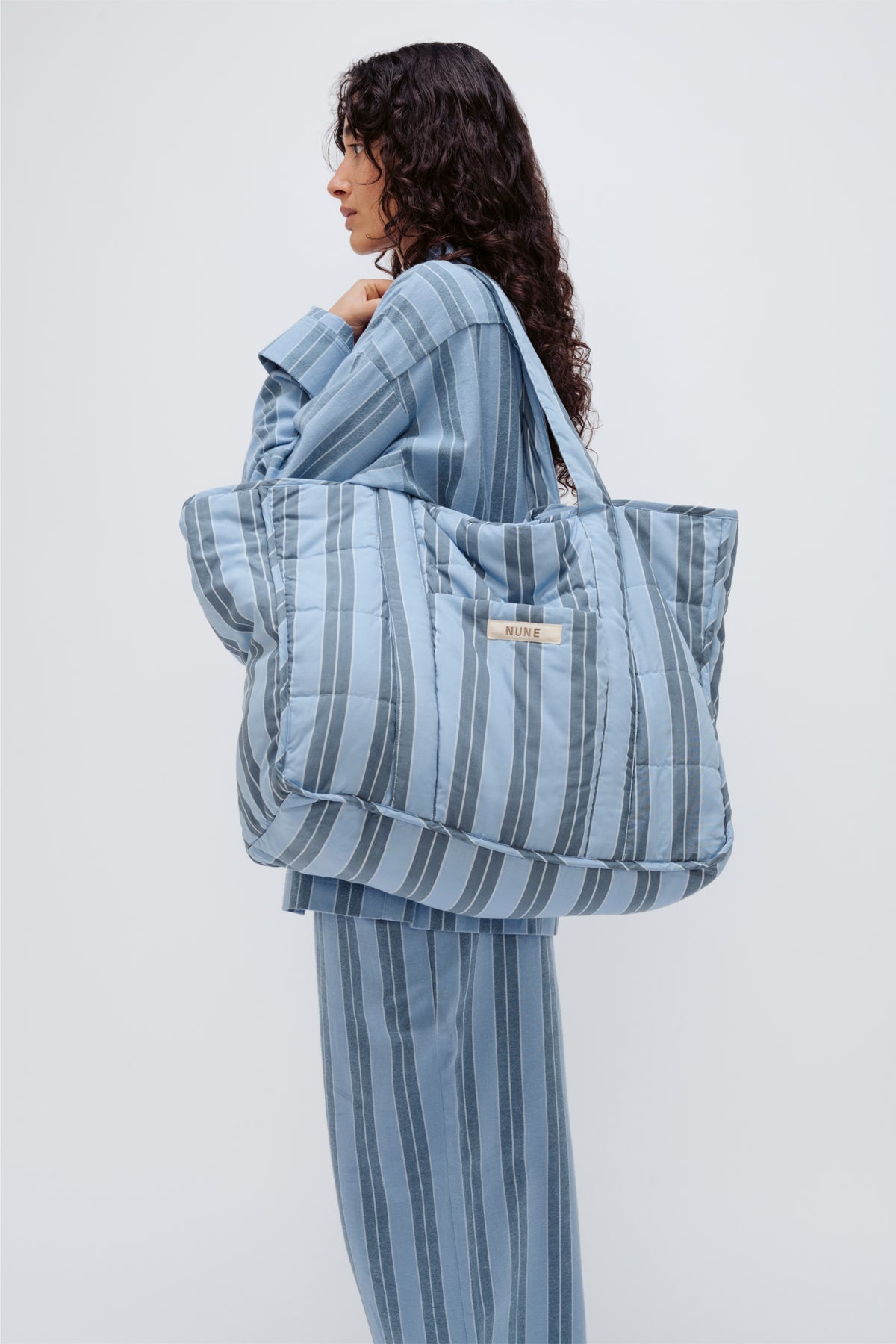 Doona Tote in Daybreak Stripe held by female model over her shoulder, wearing Long Sleeve Sleep Shirt and Long Sleep Pants Flannel in Daybreak Stripe, side view showing front pocket of Tote.