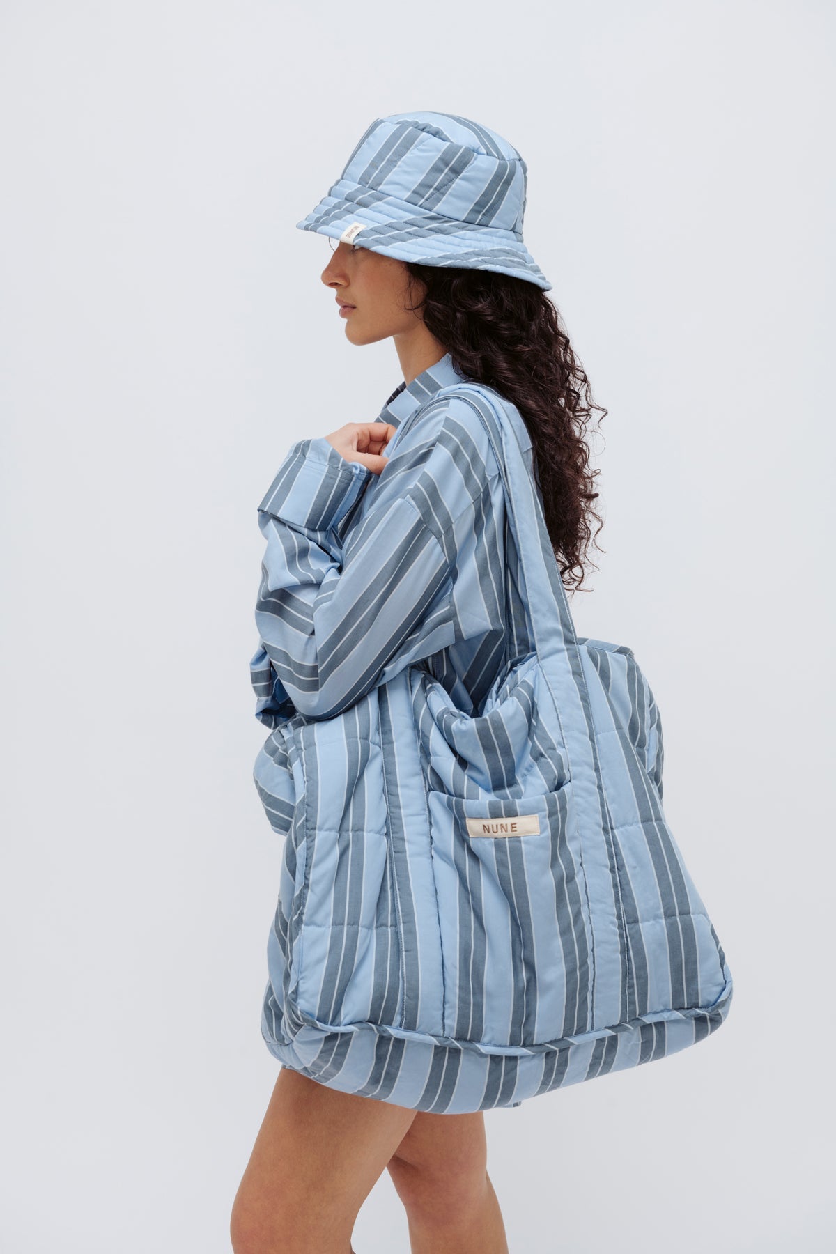 Doona Tote in Daybreak Stripe held by female model over her shoulder, wearing Long Sleeve Sleep Shirt and Doona Hat Daybreak Stripe, side view showing folded up shirt sleeve