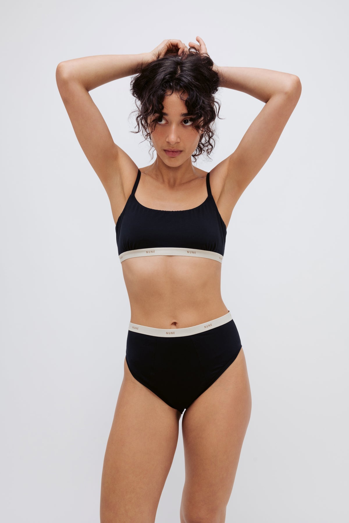 Square Neck Bralette and High Waist Brief in black worn by female model standing with arms behind her head holding her hair