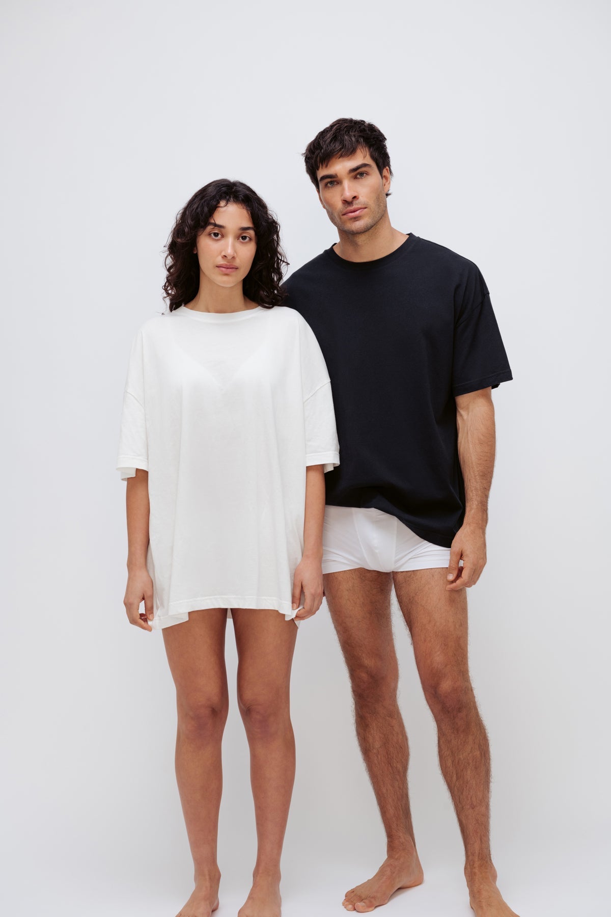 White Oversized Tee worn by female model and Black Oversized Tee with white Original Boxer Brief worn by male model, standing front view