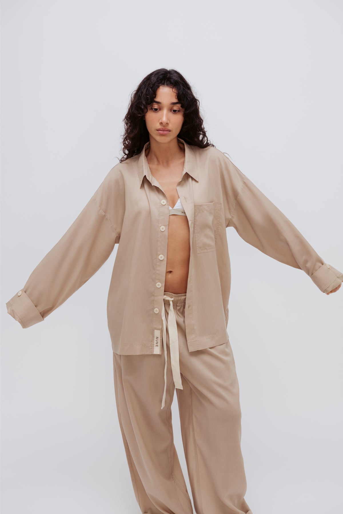 Oat long sleeve sleep shirt and long sleep pants being worn by female model over slightly visible white triangle bralette. Front view showing adjustable sleeve length on shirt and natural tie fastening on pants.