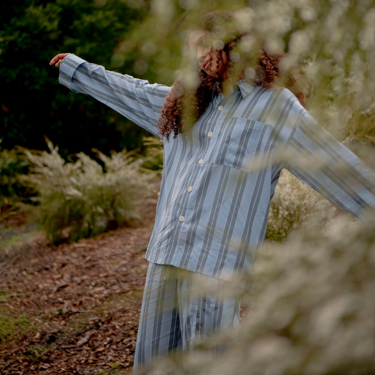 A person wearing a light blue and white vertically striped long-sleeve button-up shirt and matching pants stands outdoors in a wooded setting with blurred foliage in the foreground.