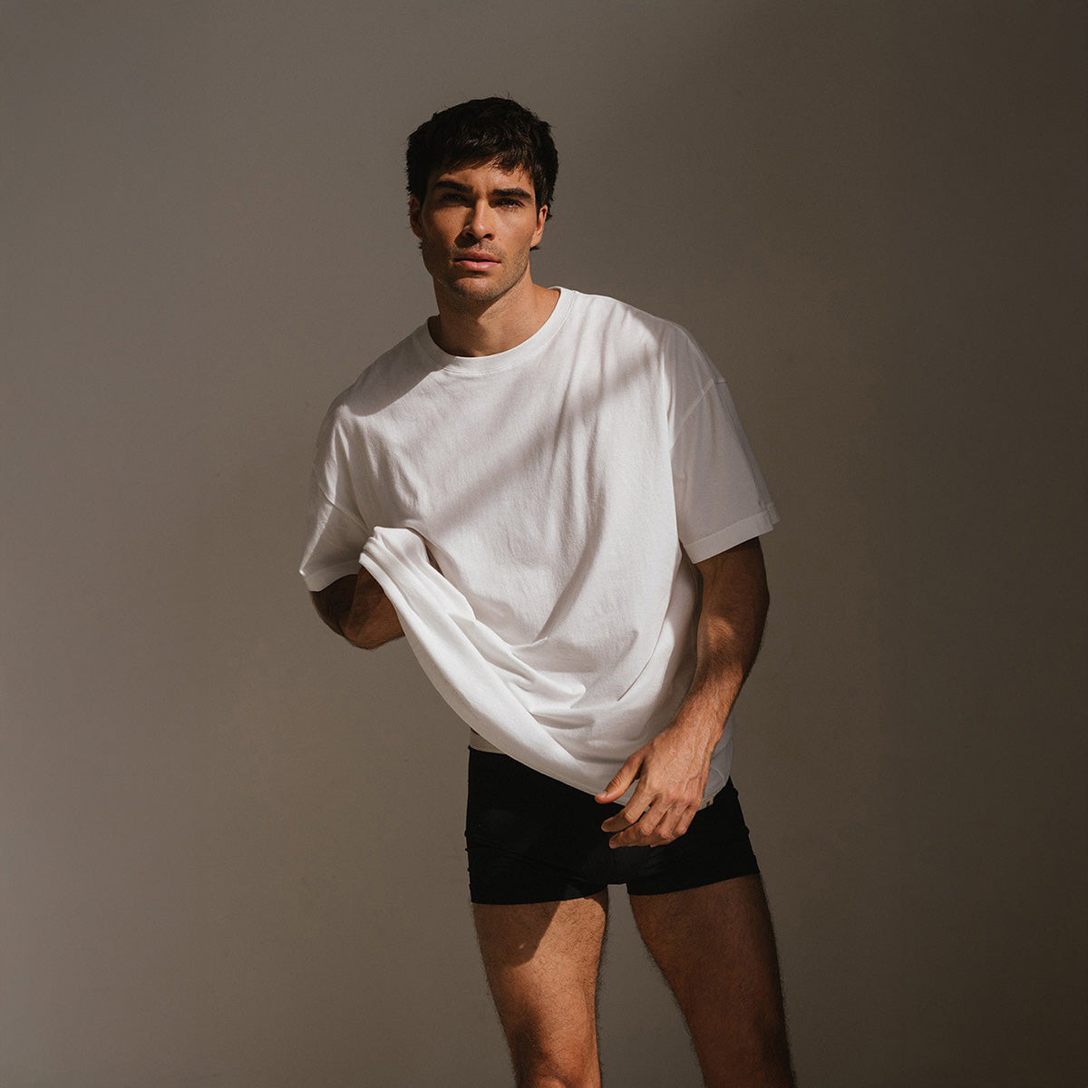 Male model wearing the NUNE Oversized Tee in White and Original Boxer Brief in Black against a warm brown backdrop.