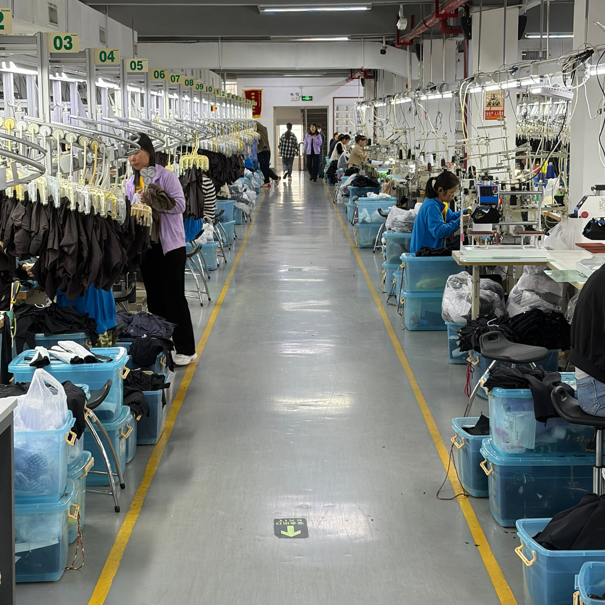 A long, narrow garment factory aisle with rows of sewing machines, workers seated and standing, and stacks of blue plastic bins filled with clothing along both sides.