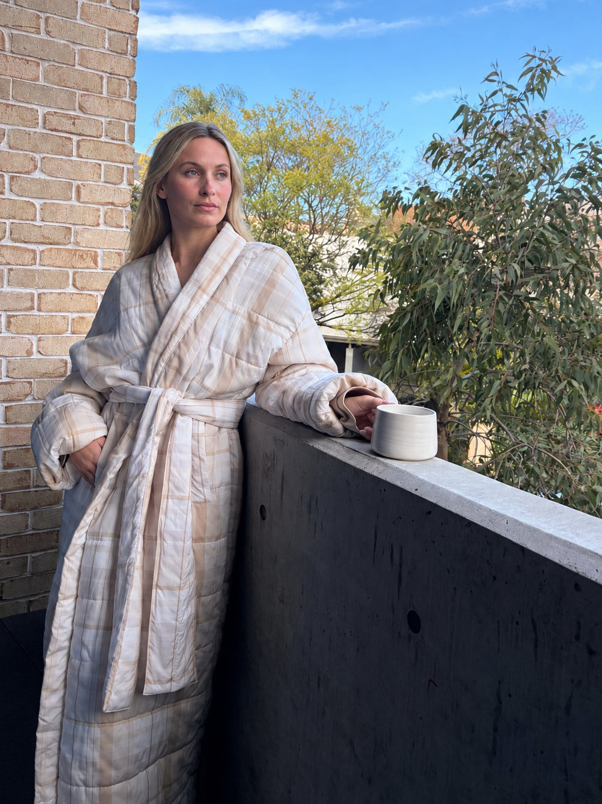 A woman wearing a cream and beige plaid quilted bathrobe stands on a balcony, leaning on a dark railing and holding a white mug. The background features a brick wall, green trees, and a bright blue sky.
