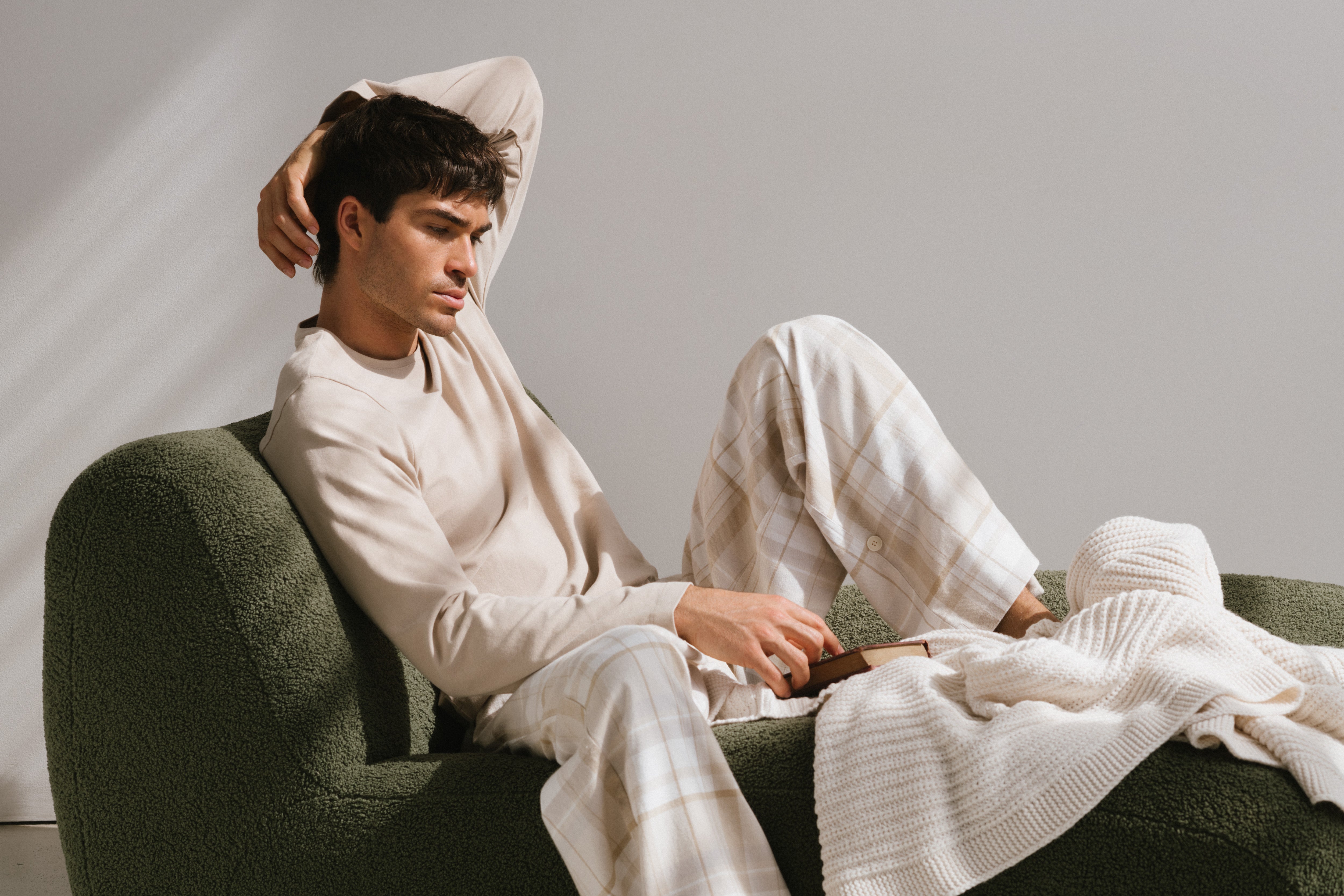 Oat long sleeve sleep top and flannel sleep pant in Oat Check worn by male model reclining on green lounge with book and cream knit blanket.