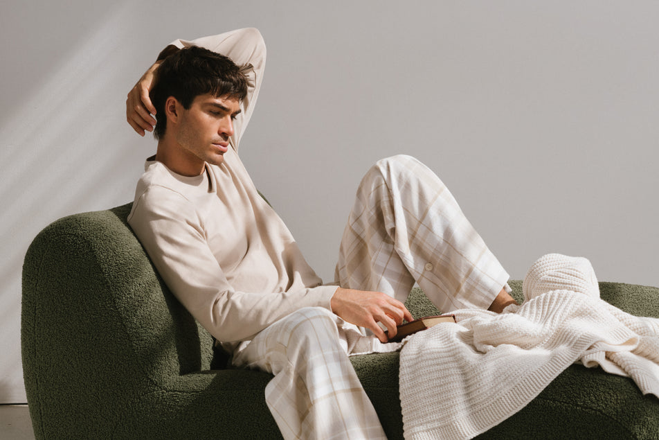 Oat long sleeve sleep top and flannel sleep pant in Oat Check worn by male model reclining on green lounge with book and cream knit blanket.