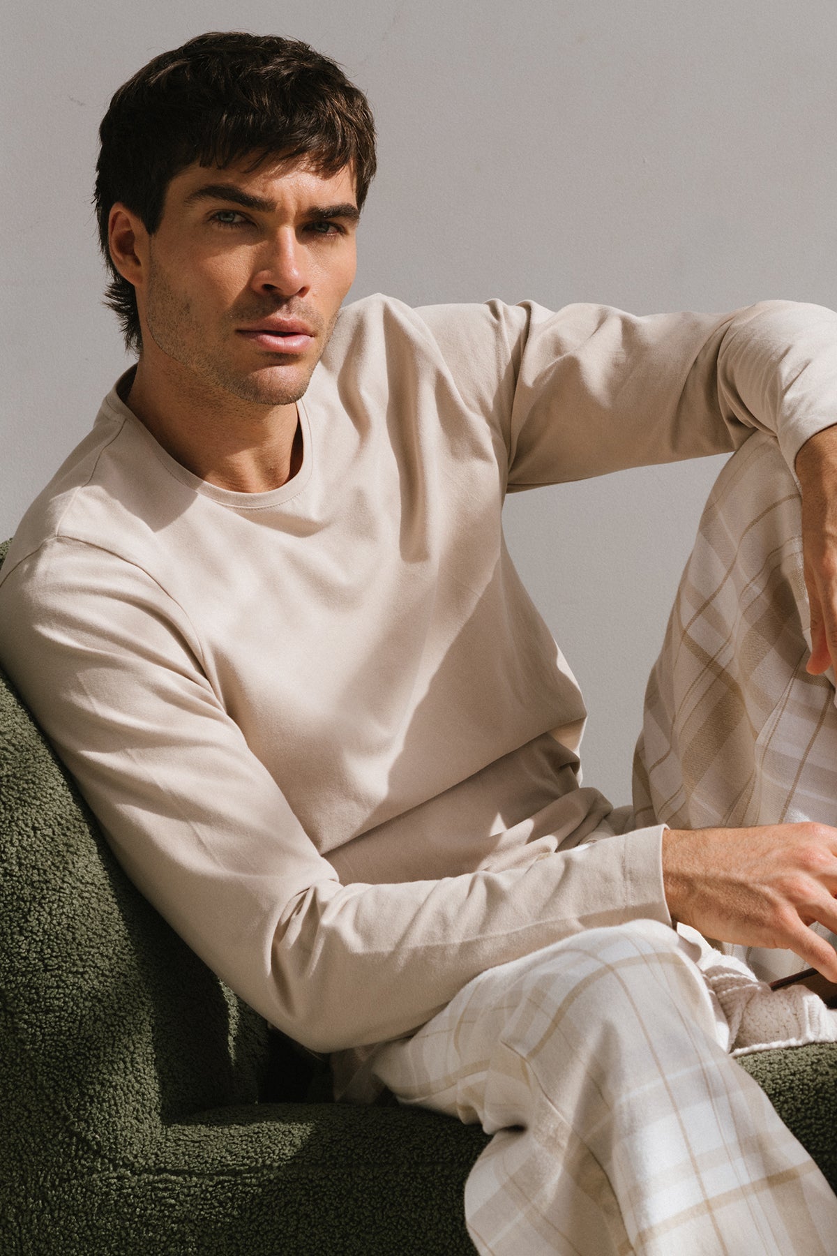 Latte Cosy Top and Flannel Sleep Pant in Oat Check worn by male model sitting on green couch in relaxed pose with arm resting on his knee 