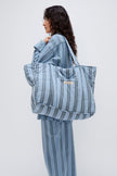 Doona Tote in Daybreak Stripe held by female model over her shoulder, wearing Long Sleeve Sleep Shirt and Long Sleep Pants Flannel in Daybreak Stripe, side view showing front pocket of Tote.