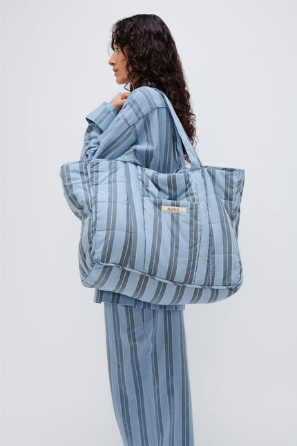 Doona Tote in Daybreak Stripe held by female model over her shoulder, wearing Long Sleeve Sleep Shirt and Long Sleep Pants Flannel in Daybreak Stripe, side view showing front pocket of Tote.