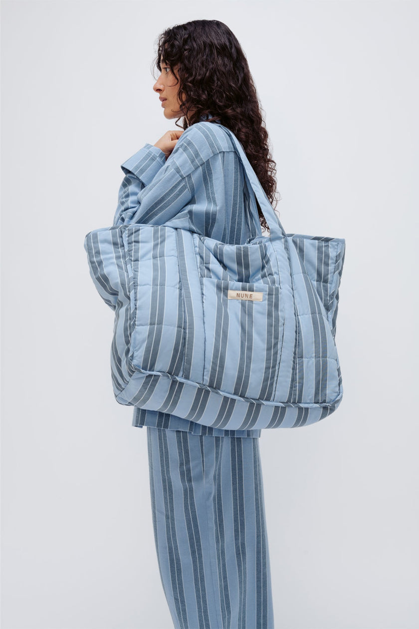 Doona Tote in Daybreak Stripe held by female model over her shoulder, wearing Long Sleeve Sleep Shirt and Long Sleep Pants Flannel in Daybreak Stripe, side view showing front pocket of Tote.