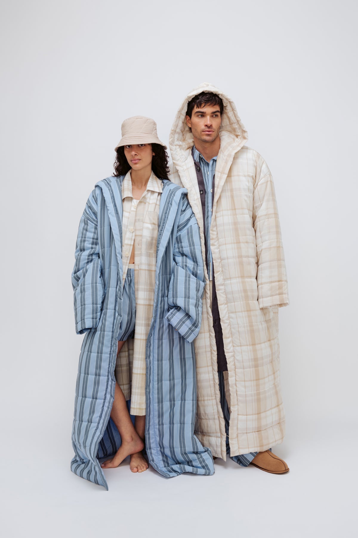 Long Doona Robe in Daybreak Stripe layered over Oat Check House Shirt worn by female model with Oat Doona Hat, and Long Doona Robe in Oat Check layered over Long Sleeve Sleep Shirt in Daybreak stripe by male model, front view with hood pulled over his head