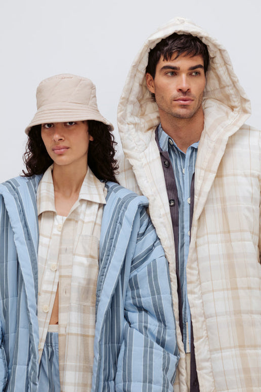 Long Doona Robe in Daybreak Stripe layered over Oat Check House Shirt worn by female model with Oat Doona Hat, and Long Doona Robe in Oat Check layered over Long Sleeve Sleep Shirt in Daybreak stripe by male model, close up front view with hood pulled over his head