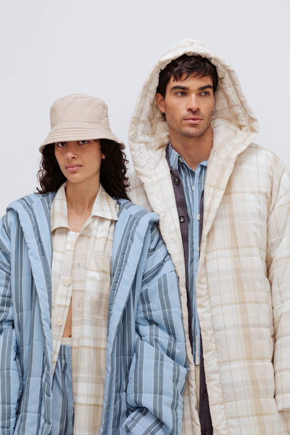Long Doona Robe in Daybreak Stripe layered over Oat Check House Shirt worn by female model with Oat Doona Hat, and Long Doona Robe in Oat Check layered over Long Sleeve Sleep Shirt in Daybreak stripe by male model, close up front view with hood pulled over his head