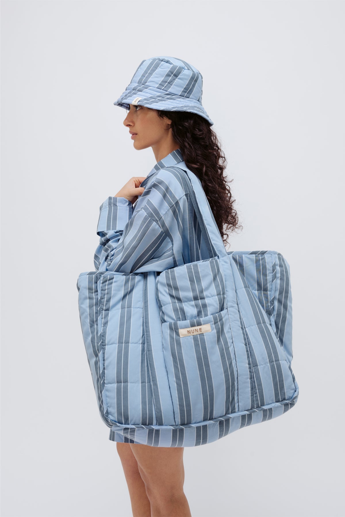 Doona Tote in Daybreak Stripe held by female model over her shoulder, wearing Long Sleeve Sleep Shirt and Doona Hat Daybreak Stripe, side view showing folded up shirt sleeve