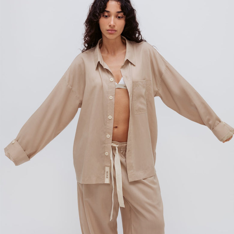 Oat long sleeve sleep shirt and long sleep pants being worn by female model over white triangle bralette.  Front view showing button fastenings on shirt and natural tie fastening on pants.