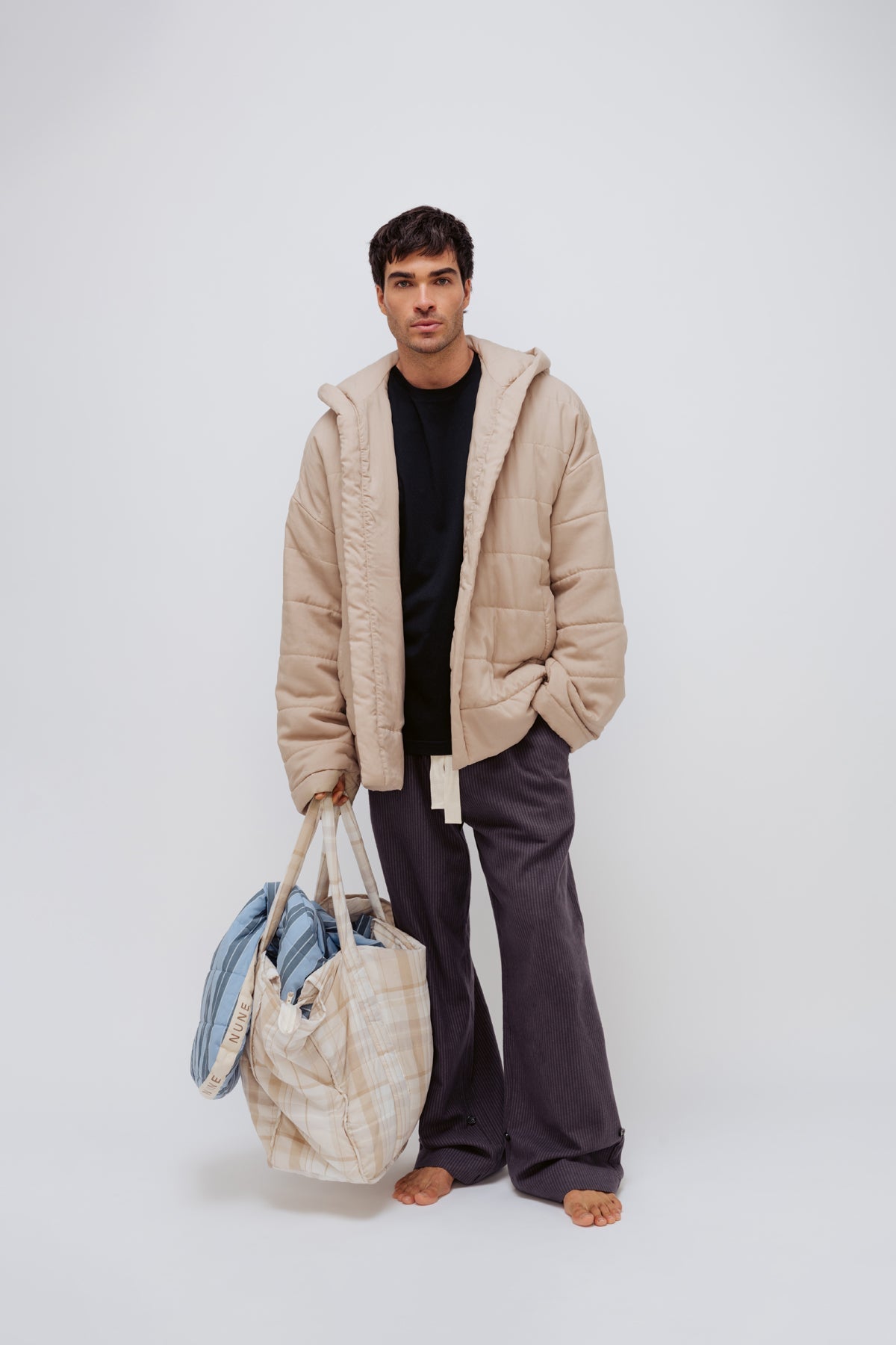 Oat short robe worn over black oversized tee and Evening Pinstripe long sleep pants by male model standing and holding the Oat Check doona tote filled with the daybreak stripe short robe