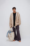 Oat short robe worn over black oversized tee and Evening Pinstripe long sleep pants by male model standing and holding the Oat Check doona tote filled with the daybreak stripe short robe