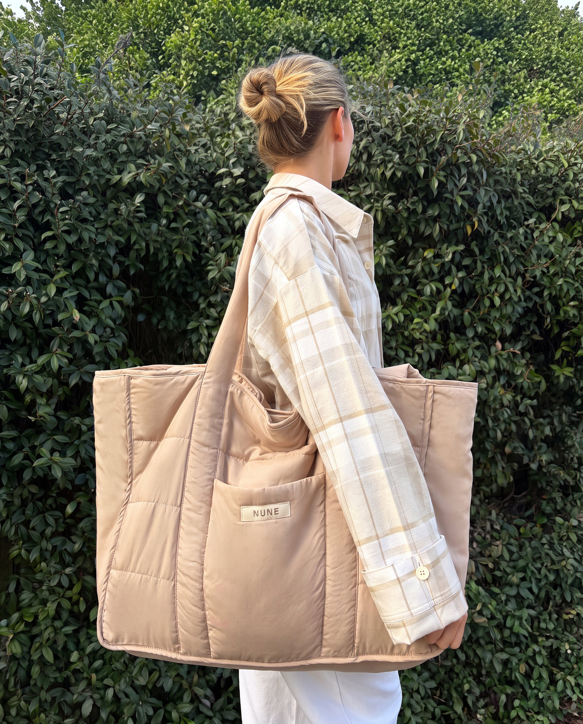 A woman in a beige plaid shirt and white pants carries a large beige puffer tote bag with a shoulder strap, standing in front of a green hedge. The bag has a small front pocket with a visible 'MINE' label.