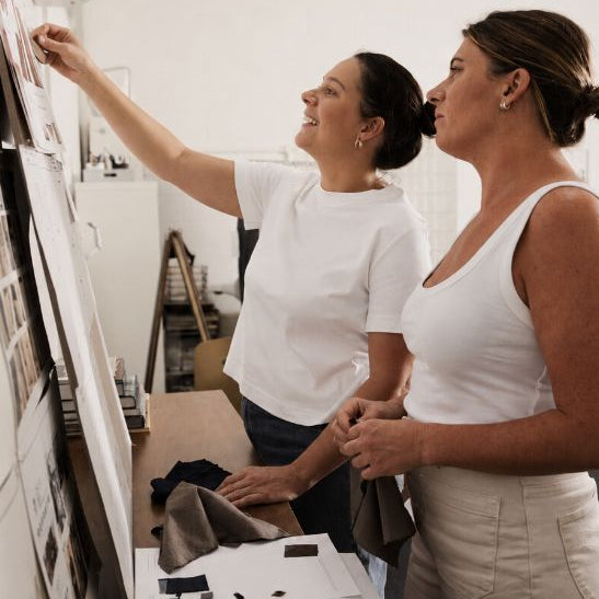 Two women in a bright, modern studio setting, looking at and pointing at a large white wall-mounted display board filled with samples and sketches. One woman wears a white t-shirt and jeans, while the other wears a white tank top and light beige pants.